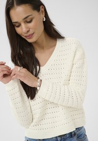 Femme aux longs cheveux bruns portant un pull en maille crème et un jean bleu, ajustant une bague en argent et un bracelet sur ses mains.