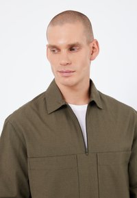 Olivengrüner Reißverschluss-Jacke mit Kragen, strukturiertem Stoff, zwei Brusttaschen und minimalistischem Design, über einem weißen Hemd.