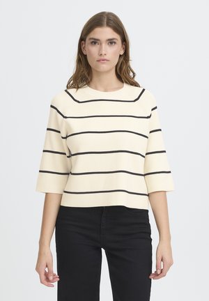 Woman with shoulder-length hair wearing a cream sweater with black horizontal stripes and black pants, standing against a plain background.