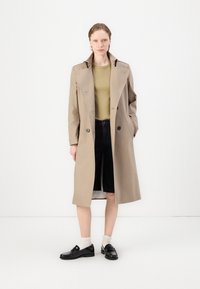 Beige trench coat with a structured design and large collar, paired with a fitted green top, black shorts, and lace-up shoes.