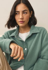 Maglia in pile verde menta, con collo a camicia, chiusura con bottoni a scatto e tessuto strutturato. Indossata sopra una maglia bianca.