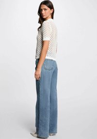 Top bianco lavorato a maglia con maniche corte e motivo aperto, abbinato a jeans a gamba larga blu chiaro a vita alta. Dettagli di cucitura sottili visibili.