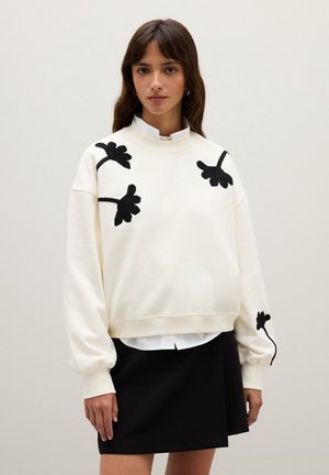 Sweatshirt blanc en tissu doux avec des broderies florales noires. Coupe décontractée avec des poignets froncés, superposé à une chemise blanche à col.