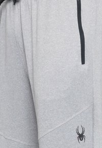 Grå sweatpants lavet af blødt stof, med en sort lynlåslomme og et lille sort edderkoplogo nær sømmet.