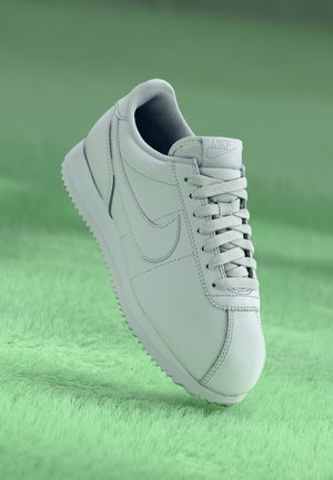 Zapatilla blanca Nike con cordones y suela acanalada sobre una superficie verde texturizada, mostrando vistas lateral y superior.