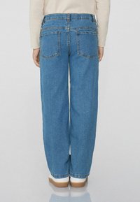 Modré rovné džínsy s vysokým pasem, dvěma zadními kapsami a jemnými detaily stehů, kombinované s bílou obuví na lemu.