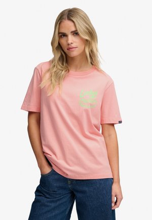 Lyshåret kvinde iført en lyserød t-shirt med grøn tekst og blå jeans, stående med den ene hånd i lommen mod en ensfarvet baggrund.