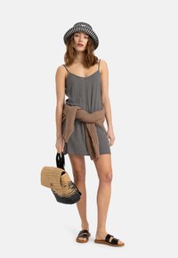 Abito a scacchi bianco e nero con spalline sottili, un maglione di maglia marrone annodato in vita, una borsa intrecciata nera e beige e sandali neri.