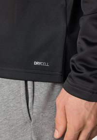 Svart sports-topp med "DRYCELL" merket på den nedre kanten, kombinert med grå joggebukser. Stoffet har en glatt, teksturert overflate.