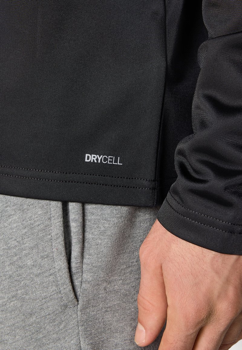 Zwarte sporttop met het label "DRYCELL" op de onderrand, gecombineerd met grijze sweats. De stof heeft een gladde, gestructureerde afwerking.