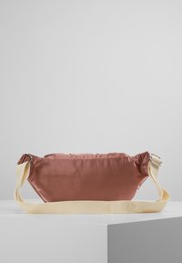 Kleine satijnen mauve heuptas met verstelbare beige stoffen riem en metalen gespen, rustend op een witte geometrische ondergrond.