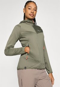 Chaqueta verde con capucha de tejido texturado, con un panel en verde oscuro en contraste, bolsillos con cremallera y tiradores de cremallera en naranja.