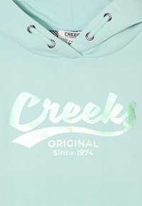 Hoodie vert menthe avec un logo holographique argenté portant la mention "Creeks Original Since 1974." Doté d'une capuche avec cordon de serrage et d'une texture lisse.
