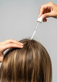 Dråbe flaske der påfører en klar væske på en opdelt sektion af langt, brunt hår med lysere striber, sat mod en lys grå baggrund.