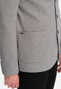 Giacca grigia a zig zag con un design strutturato, una texture sottile, una tasca sul petto e accenti di bottoni neri.