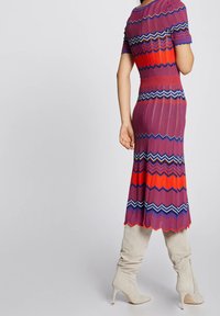 Femme portant une robe ajustée à motif zigzag rouge, violet et bleu avec des manches courtes, associée à des bottes à talons hauts beige décontractées.