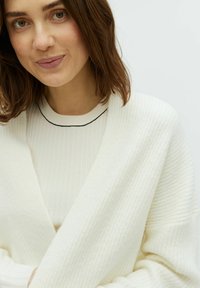 Vit ribbad cardigan med draperad framsida, bärs över en krämfärgad crew neck topp. Har en tunn svart detalj vid halsen och mjuk textur.