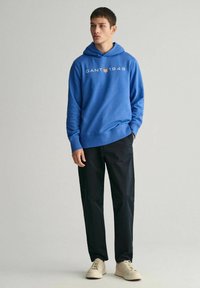 Felpa con cappuccio blu con la scritta "GANT 1949" stampata in bianco, abbinata a pantaloni neri e scarpe color crema. Stile casual, vestibilità comoda, tessuto morbido.