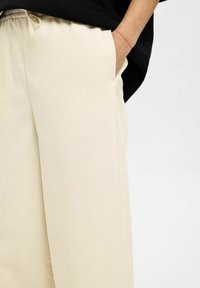 Pantalon large beige en tissu léger. Présente une taille élastique, des poches latérales et une texture douce.