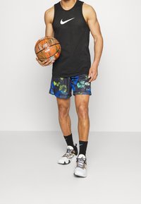 Svart Nike linne, färgglada mönstrade shorts med blå och gröna accenter, håller en basketboll, iförd vita och svarta sneakers med snören.
