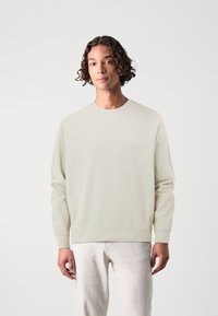 Felpa leggera color beige chiaro con girocollo in un tessuto morbido e testurizzato. Presenta polsini e orlo ribattuti. Abbinata a pantaloni chiari.