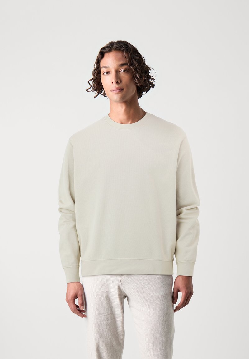 Felpa leggera color beige chiaro con girocollo in un tessuto morbido e testurizzato. Presenta polsini e orlo ribattuti. Abbinata a pantaloni chiari.