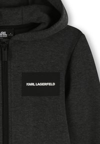 Zwarte rits-hoodie van zacht fabric, met een grote kap en een rechthoekig logopatch in het wit, geplaatst op de linkerborst.