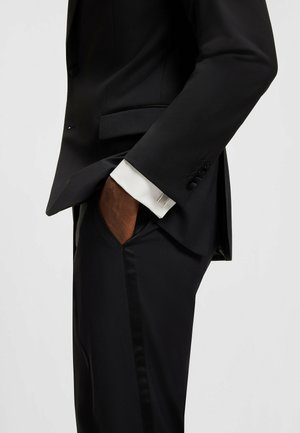 Zwarte suit met een getailleerd ontwerp, voorzien van een piek-revers, zijzakken en een satijnen strook op de broek. Witte manchet zichtbaar.