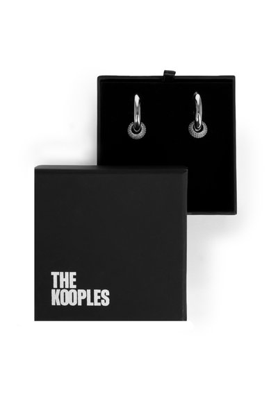 Des boucles d'oreilles en argent en forme de créoles avec un accent circulaire texturé, présentées dans une boîte noire avec un intérieur doux. Le logo "THE KOOPLES" est imprimé en blanc.