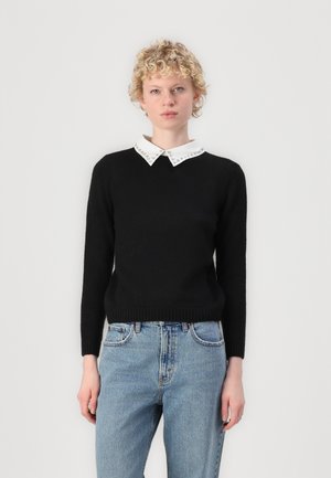 Pull noir à manches longues et col rond, avec un col blanc orné de clous décoratifs. Porté avec un jean bleu clair.
