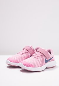 Růžové běžecké boty Nike s bílými podrážkami, síťovinovými svršky, tkaničkami a suchým zipem na jednobarevném bílém povrchu.