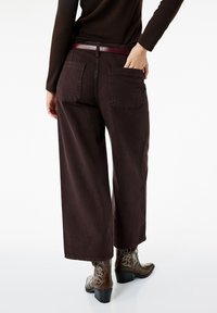 Weite, dunkelbraune Hosen mit zwei Gesäßtaschen und einem strukturierten Taillengummibund. Getragen mit braunen Stiefeletten mit Absatz, die dekorative Nähte aufweisen.