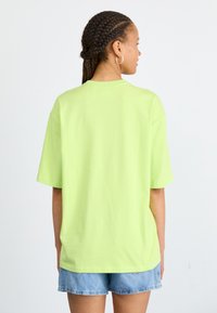 Limetkovozelené oversized tričko vyrobené z bavlny, s krátkymi rukávmi, okrúhlym výstrihom a voľným strihom, nosené s denimovými šortkami.
