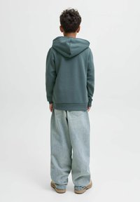 Persona in piedi di spalle, che indossa un hoodie teal, jeans azzurri larghi e scarpe beige, su uno sfondo chiaro e uniforme.