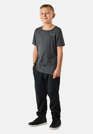 Junge steht mit den Händen in den Taschen, trägt ein dunkelgraues kurzärmeliges Hemd, schwarze Hosen und schwarze Sneaker, lächelt leicht, schlichter heller Hintergrund.