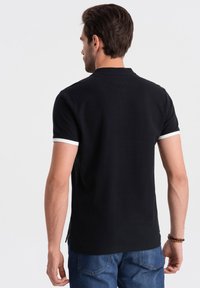 Camisa polo negra con ribetes blancos en las mangas, que presenta un diseño simple, mangas cortas y un cuello clásico, combinada con jeans azules.