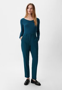 Turkusowa bluzka z długim rękawem i okrągłym dekoltem, zestawiona z dopasowanymi turkusowymi spodniami z bocznymi kieszeniami i subtelną teksturą. Całość dopełniają czarne loafersy.