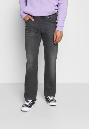 Mann trägt ausgebleichte schwarze Jeans, schwarze Sneaker mit weißen Schnürsenkeln und ein lavendelfarbenes Sweatshirt, eine Hand in der Tasche.