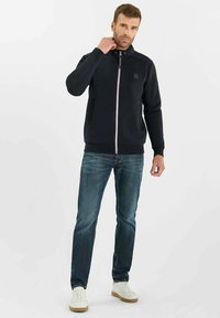 Uomo con giacca nera a zip, jeans blu e sneaker bianche, in piedi contro uno sfondo chiaro e uniforme, mentre si tocca il collo.