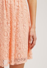 Růžové krajkové šaty s komplexními květinovými vzory, rozevlátým lemem a lehkým materiálem, doplněné přiléhavým živůtkem.