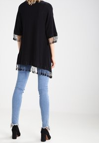 Musta tunika, jossa on lyhyet hihat ja hapsureunus helmassa. Yhdistetty vaaleansinisiin kapealahkeisiin farkkuihin ja mustiin nilkkureihin, joissa on korkea korko.