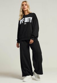 Czarna, oversize'owa bluza z napisem "OFF DUTY 1980" w białej kolorystyce, w połączeniu z pasującymi, szerokimi czarnymi spodniami oraz białymi sneakersami na platformie.