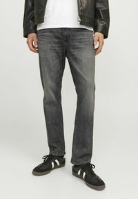 Jean en denim gris avec une coupe droite. Caractéristiques : couture visible, cinq poches et décoloration subtile. Porté avec des baskets noires et une chemise blanche.