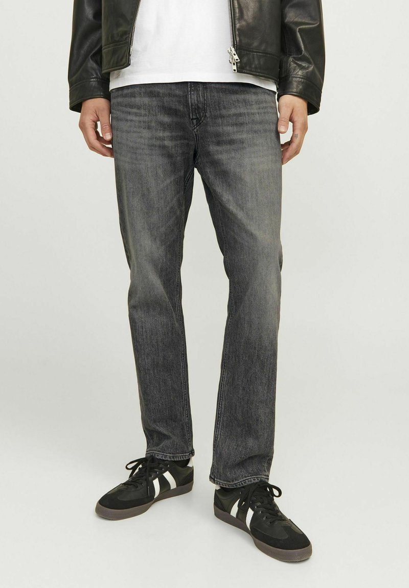 Jean en denim gris avec une coupe droite. Caractéristiques : couture visible, cinq poches et décoloration subtile. Porté avec des baskets noires et une chemise blanche.