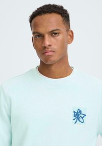 Mátově zelená mikina z měkké látky s modrým vyšívaným nášivkou na hrudi s asijsky inspirovaným textem a malým detailem kapsy.