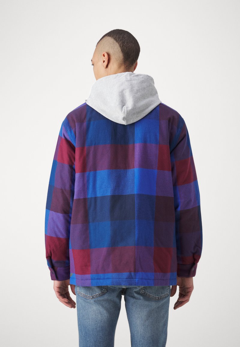 Person mit kurzen Haaren, die eine blau-rot kariertes Jacke mit einer grauen Kapuze trägt, steht mit dem Rücken zu einem schlichten weißen Hintergrund.
