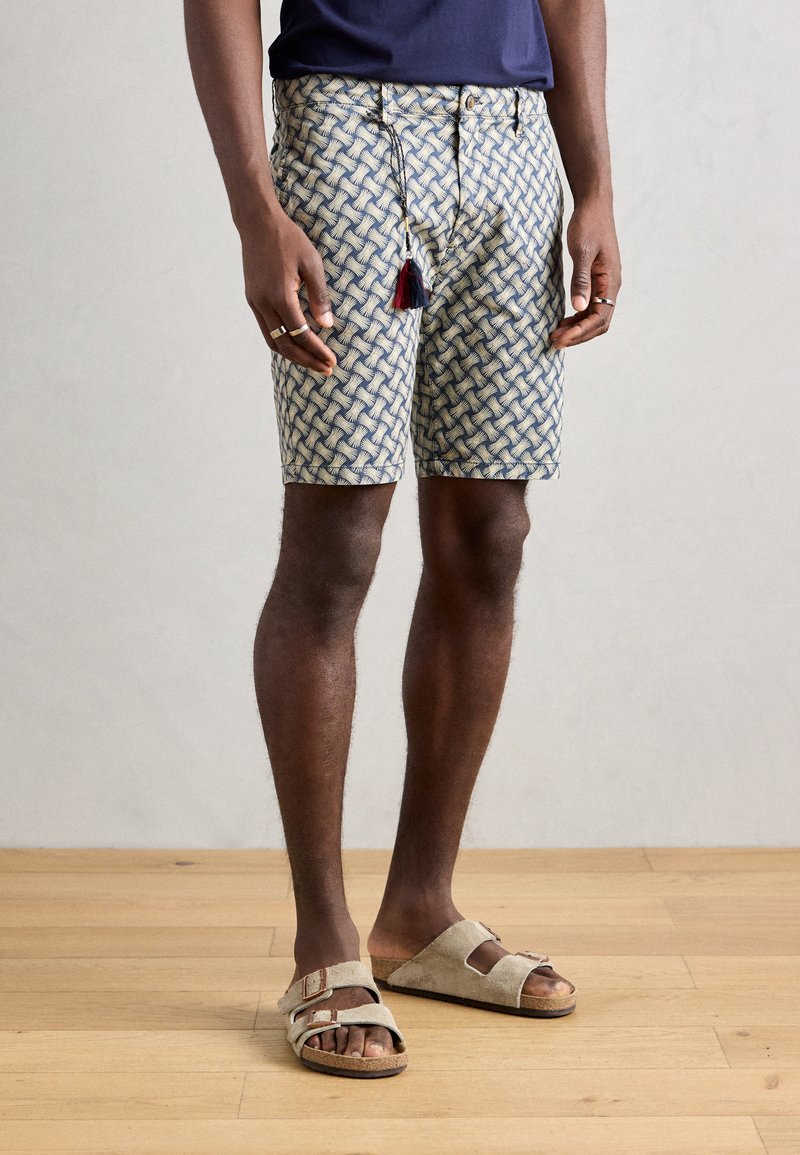 Patroonshorts in blauw en crèmekleur met een geweven ontwerp, voorzien van een trekkoordsluiting en een kwastaccent. Draagt lichtgekleurde sandalen.