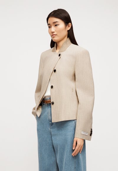 Γυναίκα που φοράει μπεζ σακάκι με κουμπιά και ψηλό γιακά, και μια ανοιχτόχρωμη μπλε τζιν καμπάνα, όρθια με το ένα χέρι στην τσέπη.