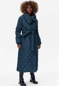 Lange gewatteerde jas in donker teal met een brede kraag, een ceintuur in de taille en een ruitpatroon. Beschikt over lange mouwen en een volledige lengte.