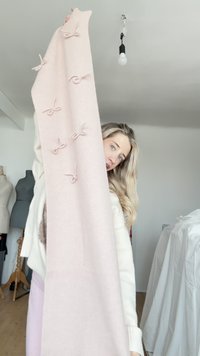 Vaaleanpunainen neulottu huivi, jossa on useita koristeellisia rusetteja sen pituudella. Pehmeä koostumus ja pitkä suorakulmainen muoto, jota henkilö pitää kädessään.